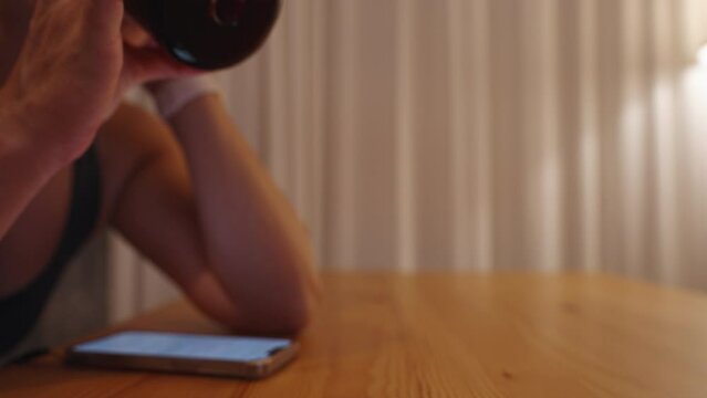 A lone young woman sits behind a wooden table, drinking whiskey with cola and looking at her phone. Dim and warm lighting comes from a lamp behind her.