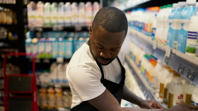 a Black man in a white t-shirt and black apron laying out bottles of milk on the counter of a supermarket with dairy products