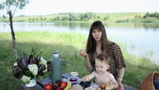 mother and little son have lunch with fruit and croissant outdoors on a wooden table against the background of the lake in summer. flowers and a thermos on the table. family weekend in nature.