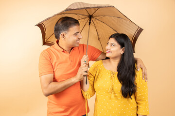 Happy indian couple standing under umbrella isolated on beige background. summer or rainy season,...