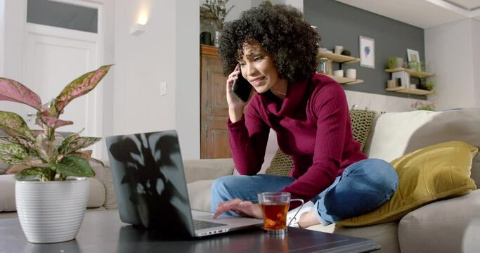 Happy biracial woman using laptop and talking on smartphone at home, slow motion