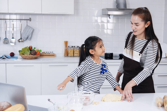 Smiling Asian mother and little asian girl child is learning online with laptop and cooking for baking bakery on wooden table together Homemade pastry for bread. Family love and Homeschool Concept.