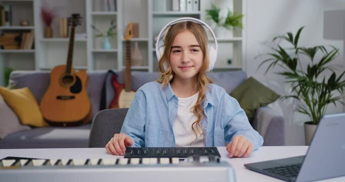 Portrait of preteen child girl learning to play music instrument and looking at camera. Beautiful little kid girl playing piano with headphones in living room or music school