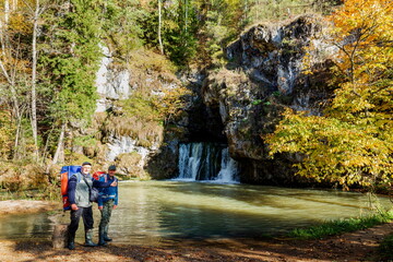Mature tourists with backpacks stand at the Atysh waterfall flowing from the Big Grotto of the Ural Mountains in the Republic of Bashkortostan on an autumn sunny day.