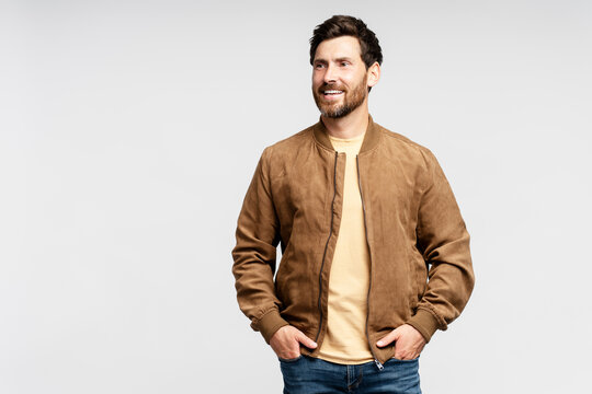 Handsome smiling bearded man wearing brown autumn jacket, stylish jeans isolated on gray background. Portrait of successful middle aged fashion model posing for pictures, studio shot 