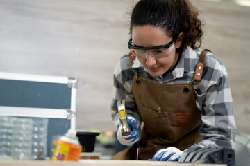 Latin woman carpenter using hammer manual tool working her wood job in carpenter's shop. Young hispanic female in protective goggles busy in furniture woodworking. Feminism in carpentry industry.