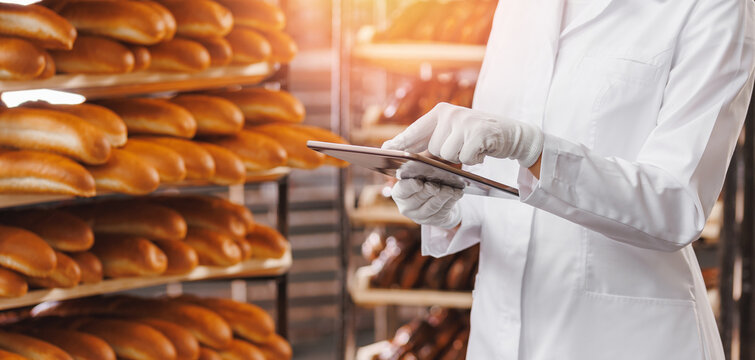 Baker woman in chef uniform use tablet computer for control quality of craft bread in bakery factory, sun light. Banner modern food industry