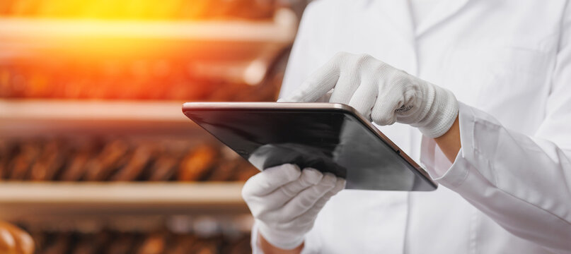 Baker woman in chef uniform use tablet computer for control quality of craft bread in bakery factory, sun light. Banner modern food industry