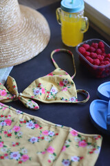 Floral swimsuit, striped towel, straw hat, cactus cup with orange soda, bowl of raspberries and blue sandals. Summer or beach essentials. Selective focus, dark background.