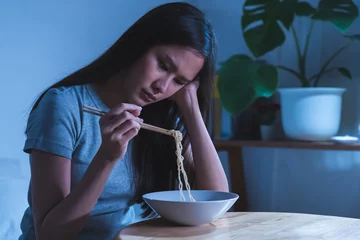 Fotobehang End of month. Alone depressed asian young student woman, girl using chopsticks eating instant ramen, noodles, cheap food and no money for dinner in bedroom in overtime night, late time at home. © KMPZZZ