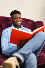 Relaxed and smiling cheerful african man on the sofa is reading a book.