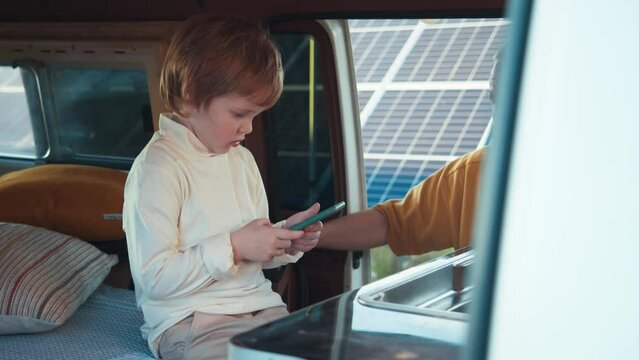 Little boy playing on smartphone, sitting inside the van. Grandfather taking care of grandson. Solar panels in the background.