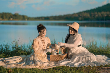Beautiful Asian woman in the Victorian or Edwardian era dressed sitting relaxed and having happy afternoon tea party in nature on the river bank, enjoying picnicking, reading books, and sipping tea.
