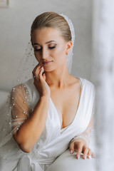 A gorgeous bride with a classic hairstyle, wearing a long robe and a long veil, is sitting and posing on a white sofa in her room. The bride on a white background