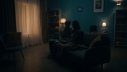 A couple sits on a sofa in a room in pitch darkness. A man holds a woman by the hand, hugs. Support for the couple, joint problem solving, reconciliation and understanding in relationships.