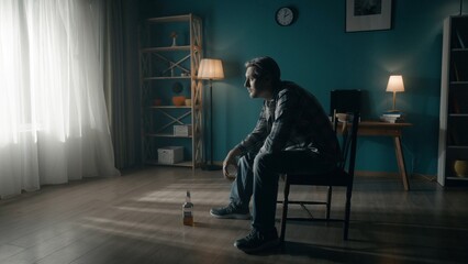 A man sits in a dark room on a chair in front of a window from which light streams. The man looks into the light, holds a glass of alcohol in his hand, there is an almost empty bottle on the floor.