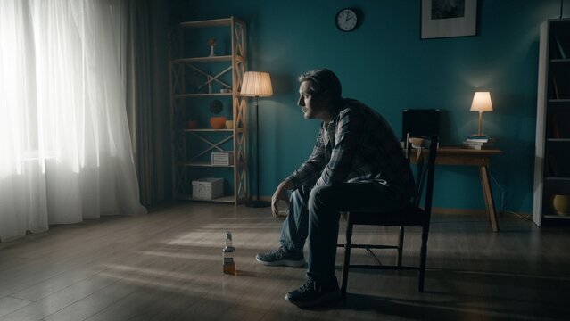 A man sits in a dark room on a chair in front of a window from which light streams. The man looks into the light, holds a glass of alcohol in his hand, there is an almost empty bottle on the floor.
