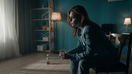 A woman sits in a dark room on a chair close up, in front of a window. A woman looks into the light, holds a glass of alcohol in his hand, there is an almost empty bottle on the floor.