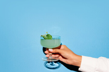 Female hand holding glass with cocktail on bright pop art blue background with shadow. Copy space for ad.