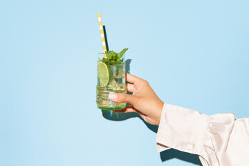Lady's hand holding glass of mojito with lime on light blue background.