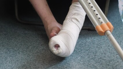 a broken leg in a cast in a bandage of a young man sitting with crutches. consequences of fractures and injuries, medical insurance, temporary disability