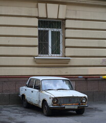 An old rusty white Soviet car stands under the window of a residential building, Sedova Street,...