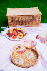 Fototapeta na wymiar Pink drinks, cocktail with ice, raspberry, rosemary. Two glasses with martini, champagne, cider, lemonade on the blanket with fruit plate, picnic basket. Cozy summer picnic on nature. Selective focus.