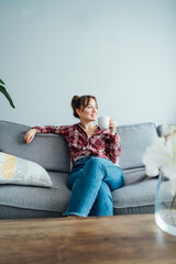 Young smiling woman sitting on sofa and looking side up while drinking coffee. Young brunette woman...