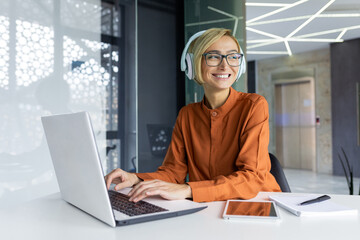 Young beautiful woman working on laptop inside office at workplace, smiling businesswoman listening...