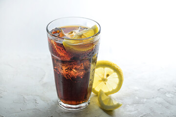 Fresh cola with ice cubes and lemon slices in a drinking glass, sweet caffeine drink against a light gray background, copy space, selected focus