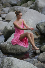 Portrait of beautiful young woman in bright pink dress posing on the large stones in the riverbed. Concept of wellbeing, mindfulness, connecting with nature and enjoying the healing sound of water