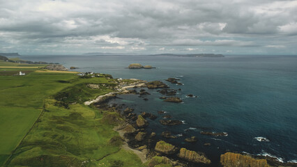 Rocky shore aerial view: green grass meadows and fields. Water surface stretches to horizon with grey clouds. Alone building on cliff. Northern Ireland panoramic landscape.