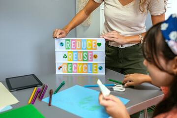 Unrecognizable female teacher showing lightbox with text reduce, reuse, recycle while student drawing on colorful cardboards in ecology classroom. Sustainable lifestyle education concept.
