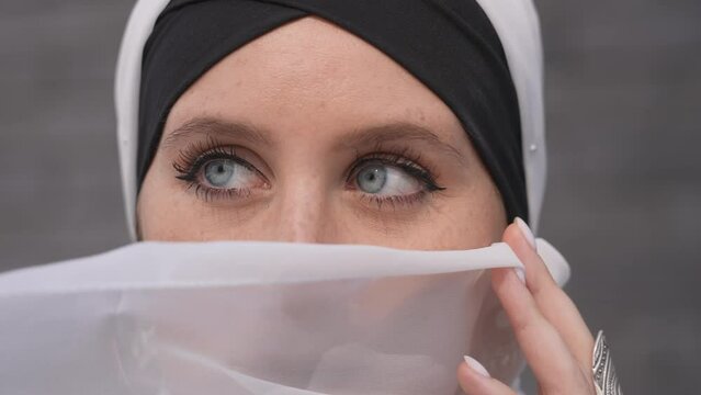 Portrait of a young blue-eyed woman in a hijab against a gray brick wall. Muslim woman with white scarf covering her face. 