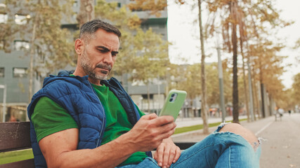Mature man tourist wearing casual clothes sitting on bench scrolling smartphone