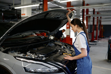 Young woman car mechanic looking under hood while working at repair garage