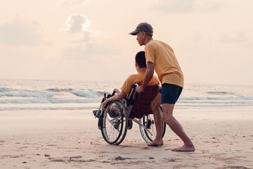 Fototapeta premium A parent, caregiver or volunteer helping teenage boy push wheelchair to the beach in evening sunset,exercise and learning about nature on the sea,Travel on vacation with family, Mental health concept.