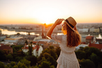 Fototapeta premium Happy traveler in a stylish dress and hat enjoys the sunrise or sunset with stunning views of the city. Back view. Lifestyle, travel, tourism, nature, active lifestyle.