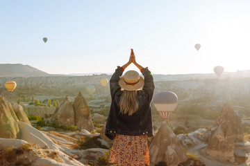 Attractive young woman in hat stands on mountain with her back, enjoying view of flying hot air balloons. Enjoy the moment. Concept of freedom, active lifestyle. Famous tourist region Cappadocia.