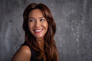Close-up of a beautiful brown-haired woman standing in front of a grey wall and cheerful smiling