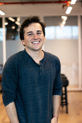 young white latino man with brown hair and in a trendy gray shirt smiling towards the camera happily in a warm modern workspace