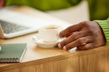 Focus on male hand taking coffee cup. Desk for work with laptop and notebook on it. Business atmosphere. Concept of business and education, freelance job, modern lifestyle