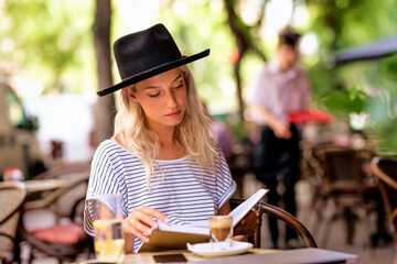 Blond haired woman wearing hat and reading a book on the outdoor cafe terrace