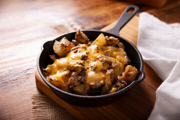 Homemade recipe for potatoes with steak and melted cheese, seasoned and served in a cast iron skillet, on a rustic wooden table.