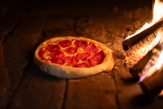 Italian pizza pepperoni is cooked in a wood-fired oven. Pizza with salami in hot oven near burning firewoods close up. Food photography