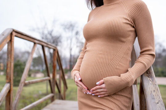 Close-up Pregnant beautiful girl in a beige tight-fitting dress stands near the wooden stairs in the park, holding her hands on her stomach. Side view
