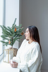 Woman in warm white winter sweater standing next to the window at home at christmas eve holding cup with marshmallows, fir tree behind