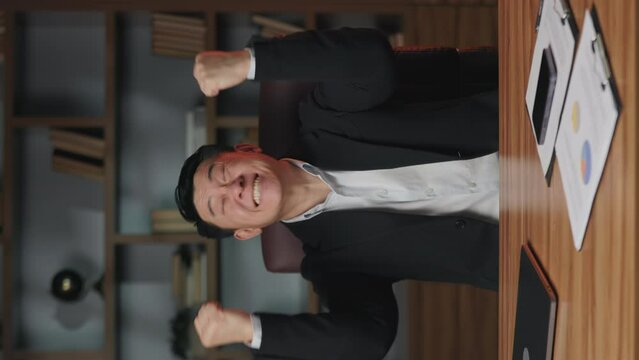 Close up view of emotional entrepreneur in business suit lifting fists in winner gesture while staying indoors. Vertical video of thrilled employer screaming in excitement while sitting in office.