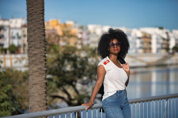 Young, beautiful, black woman with afro hair, white t-shirt and sunglasses leaning on a metal railing, on vacation, receiving the sun's rays. Concept travel, relax, vacation, current, modern.