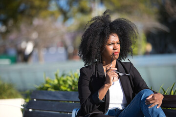 Fototapeta na wymiar Young, beautiful, black woman with afro hair, wearing a jacket, with the temple of her glasses on her mouth, pensive, sitting on a bench. Concept thoughts, dreams, doubts, current, modern.